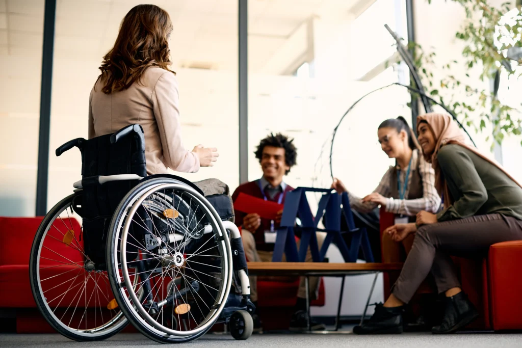 Vista general de una reunión en donde se puede ver el fondo desenfocado y se puede observar a una mujer feliz en su silla de ruedas.
